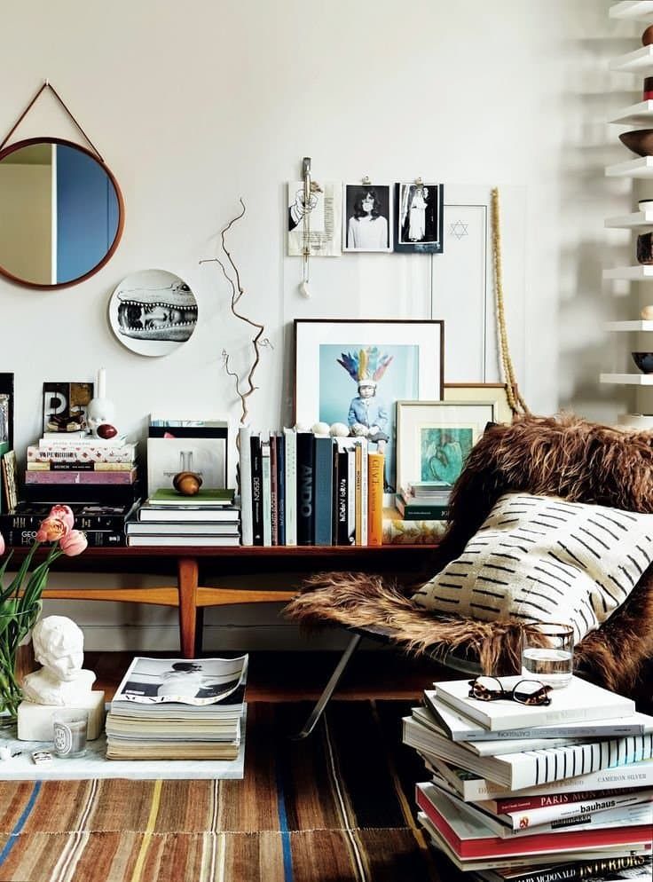 A detail shot of a bohemian-inspired reading corner featuring a mid-century wooden bench overflowing with stacked books, artwork leaning against the wall, and dried branches in a vase. A furry chair with patterned pillows invites relaxation, surrounded by more book stacks and personal touches like a small bust and a round mirror.