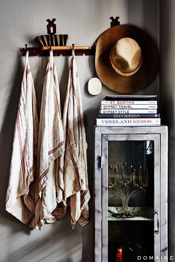 Charming hallway nook featuring textured linen towels with red stripes hanging on a weathered rack. A wide-brimmed hat and a collection of books add a touch of lived-in comfort above a glass-doored cabinet.