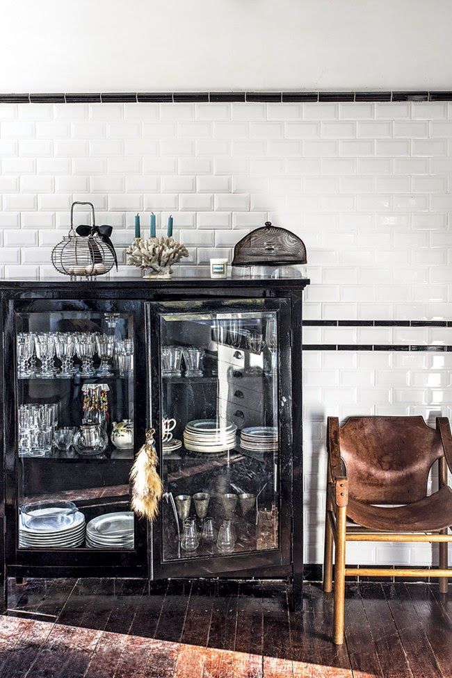 A dark, possibly antique, cabinet with glass doors showcases various glasses, jars, and stacks of white plates. On top of the cabinet are a black wire basket, a coral-shaped candle holder with blue candles, and a wire mesh dome. To the right of the cabinet is a brown leather sling chair with a wooden frame. The background features white subway tile walls with black trim and a dark wooden floor with sunlight streaming in.