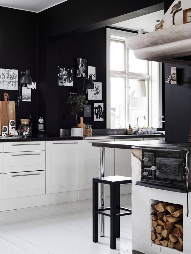 A striking kitchen interior with matte black walls contrasting with clean white shaker-style base cabinets. A black countertop holds various kitchen items and decor. In the foreground, an antique black cast iron wood stove with a white base and visible firewood adds a rustic touch. A large window with white trim is visible in the background, and small framed prints adorn the black wall.