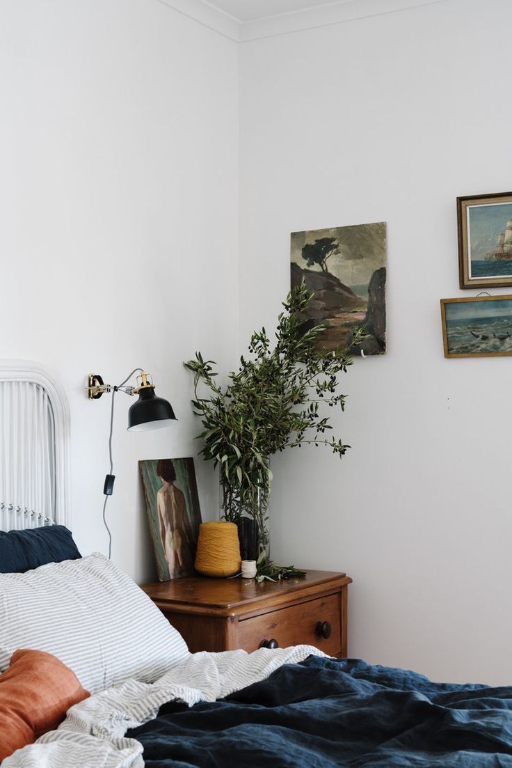 A bright bedroom corner showcasing natural textures, including a bed with a white rattan headboard and deep blue linen bedding. A wooden nightstand holds a clear vase filled with leafy branches, a mustard yellow yarn spool, and a small framed painting. A black wall-mounted lamp is above the nightstand, and several framed landscape paintings hang on the white wall.
