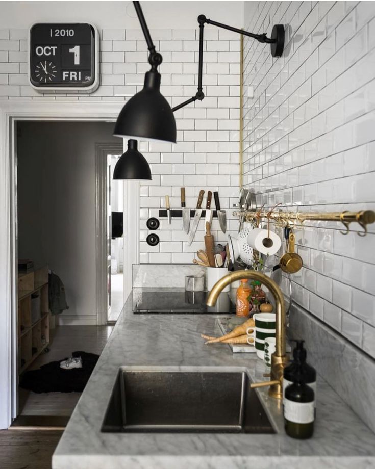 A kitchen featuring white subway tile walls, light gray marble countertops with an undermount sink, and a prominent brass faucet. Black industrial-style sconces illuminate the space, and kitchen tools hang from a brass rail on the wall.