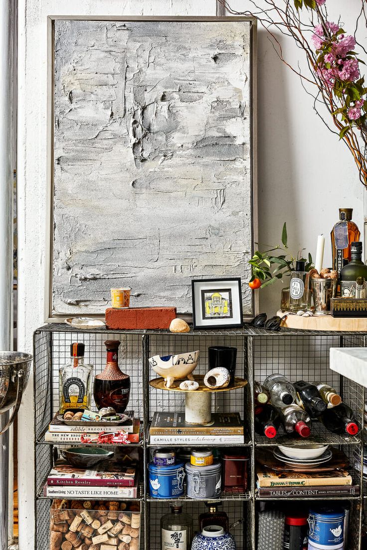 A wire shelving unit in a boho-style kitchen, displaying a mix of liquor bottles, cookbooks, decorative objects, and a wine rack, topped with an abstract grey painting and various styled items.