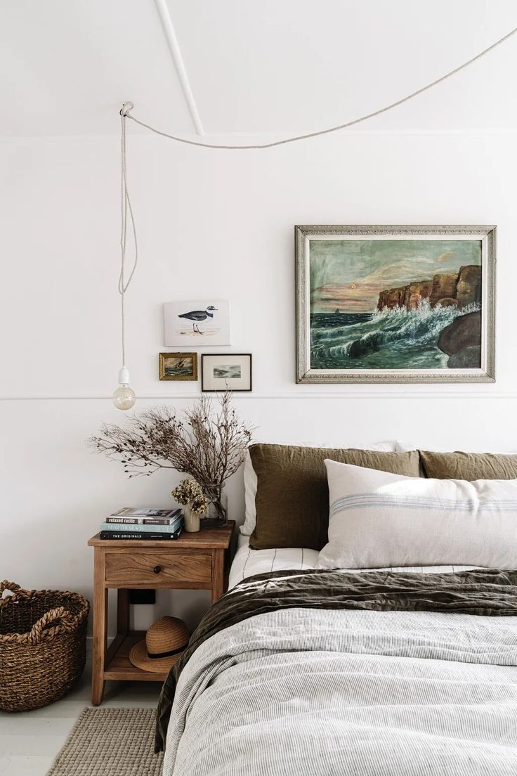 A cozy bedroom corner with a natural wood nightstand holding a stack of books, a vase with dried branches, and a straw hat. The bed is made with olive green and striped linens, and a large framed seascape painting hangs above the headboard, flanked by smaller bird and landscape prints. A bare bulb pendant light hangs from the ceiling.