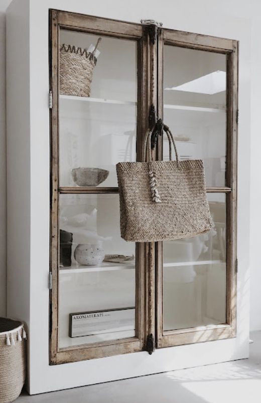 A white display cabinet with distressed wooden frames on its double glass doors. A woven straw tote bag with braided handles and tassels hangs on a hook from the right door. Inside the cabinet, various light-colored ceramic bowls, a woven basket, and other decorative items are visible on shelves.