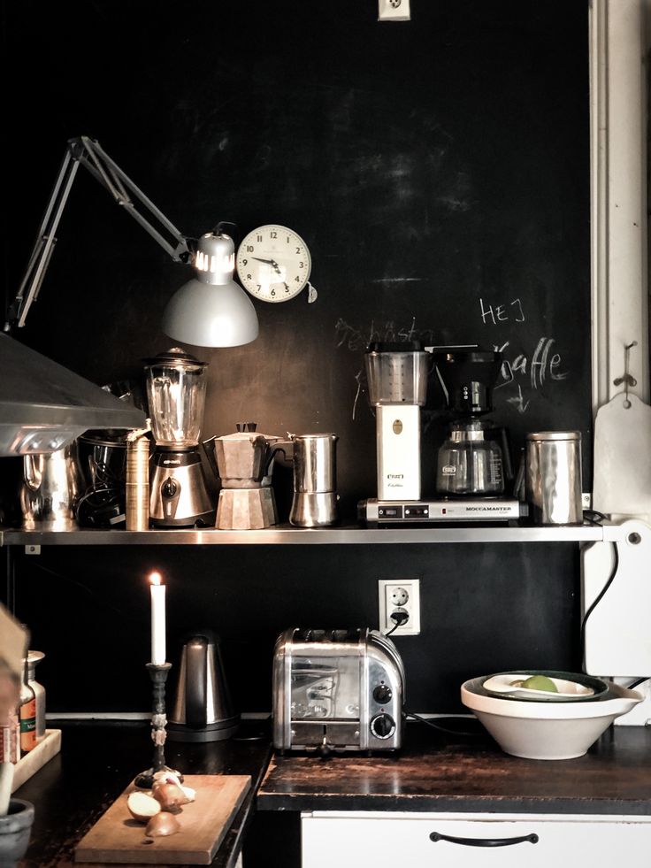 A close-up view of a kitchen counter and an open shelf set against a distressed black chalkboard wall. The top shelf holds multiple coffee appliances including a percolator, a blender, and drip coffee makers, along with metal containers. A silver desk lamp and a round white clock are mounted on the wall. Below, on the counter, there's a lit white taper candle in a dark holder, a chrome toaster, a cutting board with onions, and a white bowl. Some writing is visible on the chalkboard wall.
