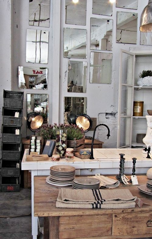 A rustic and eclectic kitchen or retail space featuring a wall covered with an arrangement of antique-style mirrors, a farmhouse sink, stacks of plates, and vintage metal crates.