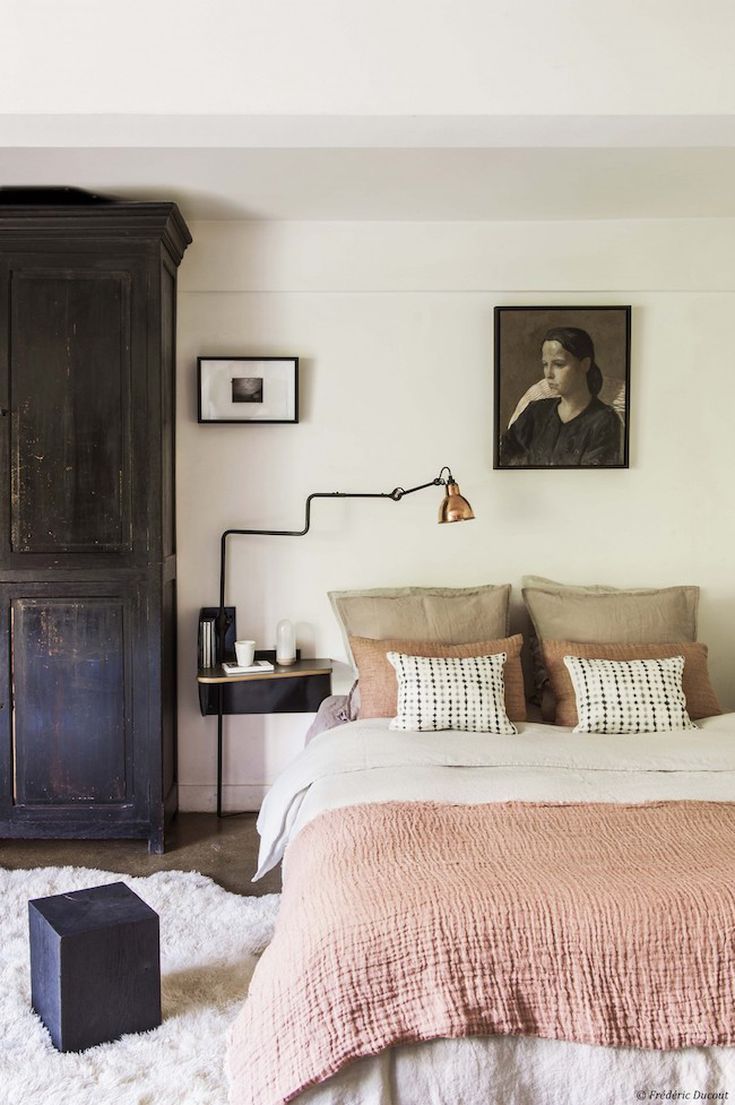 A bedroom designed with natural textures, showcasing a large, dark distressed wood armoire next to a bed dressed in soft pink, white, and neutral-toned linen and cotton bedding. A fluffy white rug covers the floor, and a black wall-mounted nightstand with a bronze-accented lamp is beside the bed. Artwork hangs on the light-colored wall.