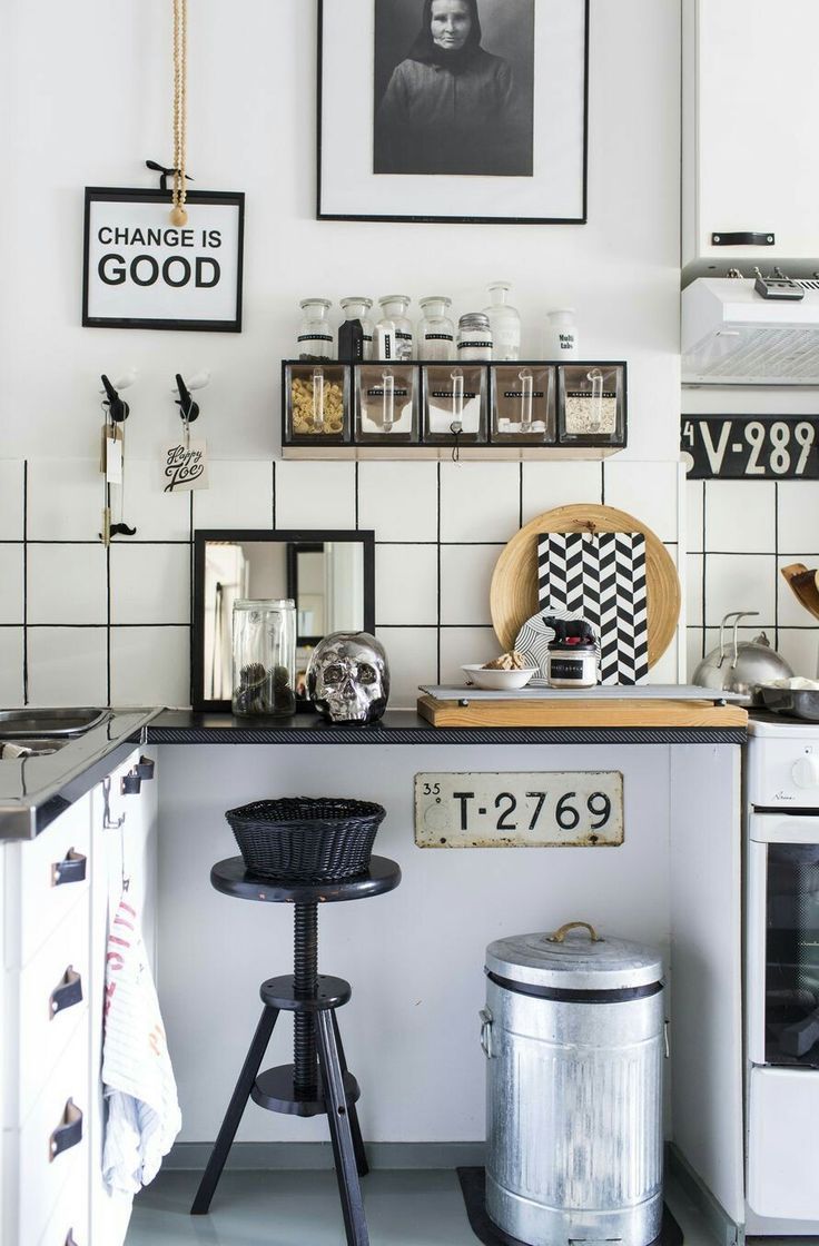 An eclectic kitchen counter area showcasing white square wall tiles with dark grout. On the black countertop, a decorative silver skull, a small black mirror, and a wooden tray with a black and white chevron board are displayed. Above the counter, a black shelf holds clear jars of dry goods. A vintage-style license plate with "T-2769" is affixed to the counter's base, and a tall black stool holds a woven basket. A large metal trash can sits on the floor.