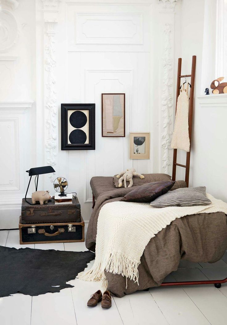 A cozy, eclectic bedroom featuring a bed with brown linens and a cream fringed throw, a unique nightstand made of stacked vintage suitcases holding a black desk lamp and small fan, and a lean-to ladder acting as a clothing rack. Art in black frames hangs on the ornate white wall, and a dark animal-hide rug is on the white floor.