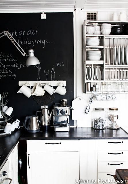 A cozy kitchen corner featuring a black chalkboard wall with white writing, white hanging mugs, a coffee station on a black countertop, and white cabinets.