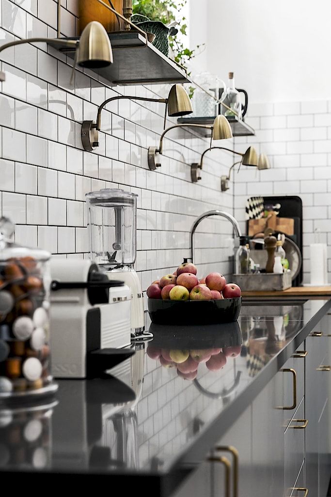A modern kitchen featuring a white subway tile backsplash, sleek dark gray countertops reflecting the light, and several arched brass wall sconces providing task lighting above. A bowl of apples and a coffee machine sit on the counter.