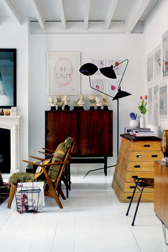 Eclectic interior design showcasing a mid-century modern aesthetic with a dark wood credenza, abstract art, two striped armchairs, and a stack of vintage wooden gym boxes serving as a side table in a room with white walls and floors.