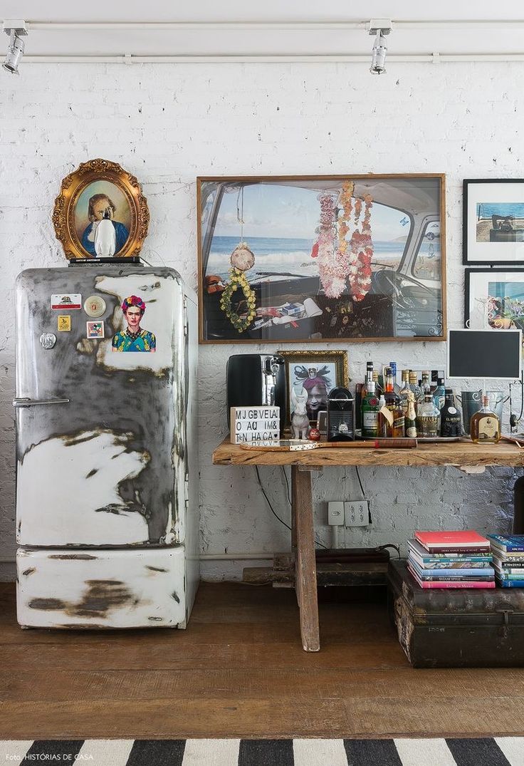 Unique and bohemian interior with exposed white brick walls, a heavily patinated vintage refrigerator topped with a framed portrait, a large, colorful artwork showing flowers in a car, and a distressed wooden bar table.