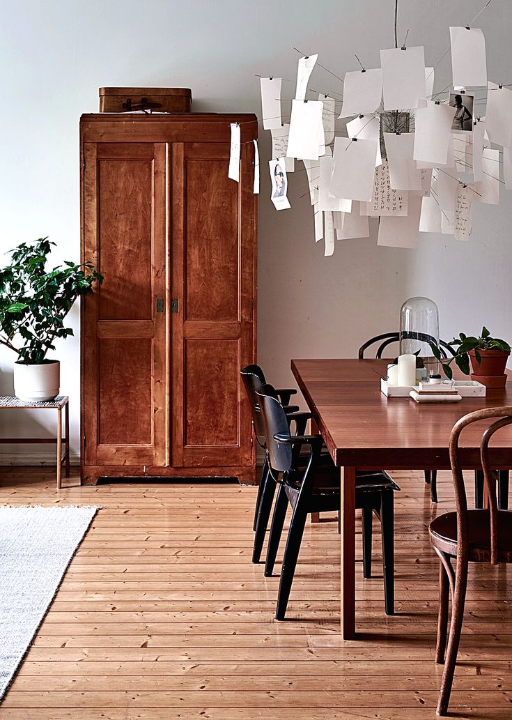A minimalist dining space with warm wooden floors, a large antique wooden armoire, and a unique DIY chandelier crafted from numerous hanging papers. A potted plant sits on a metal stand.