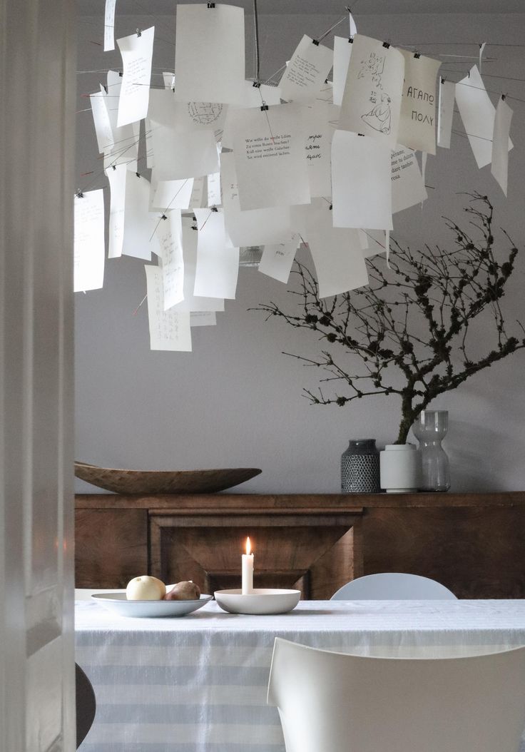 An artistic chandelier crafted from numerous small, white papers clipped to wires, illuminating a dining room. Below it, a dark wood credenza holds a dried branch arrangement, and a white tablecloth covers a table with a lit candle.