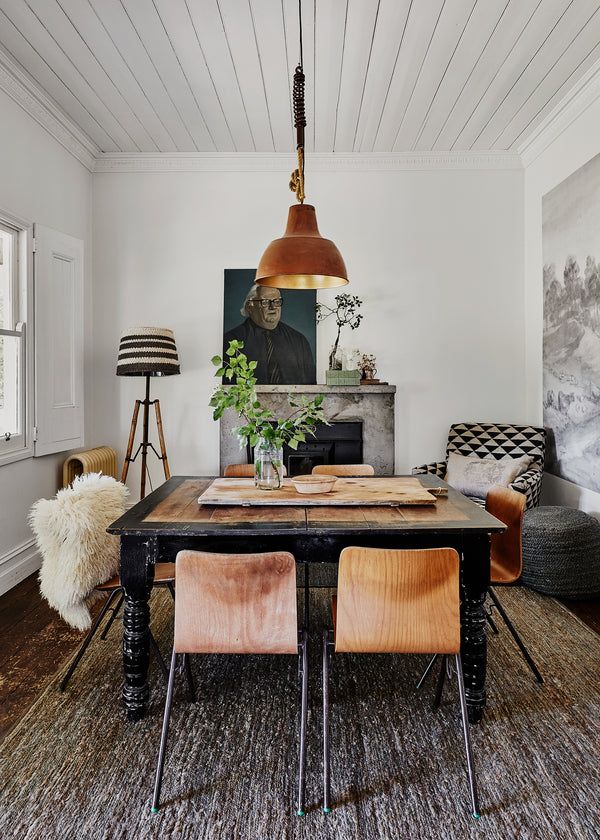 A dining area with a heavy black base wooden table and four wooden chairs with metal legs, set on a textured rug. Overhead hangs a large industrial-style copper pendant light. In the background, a dark portrait hangs above a rustic fireplace, flanked by a tall, striped floor lamp and an armchair with a geometric pattern.