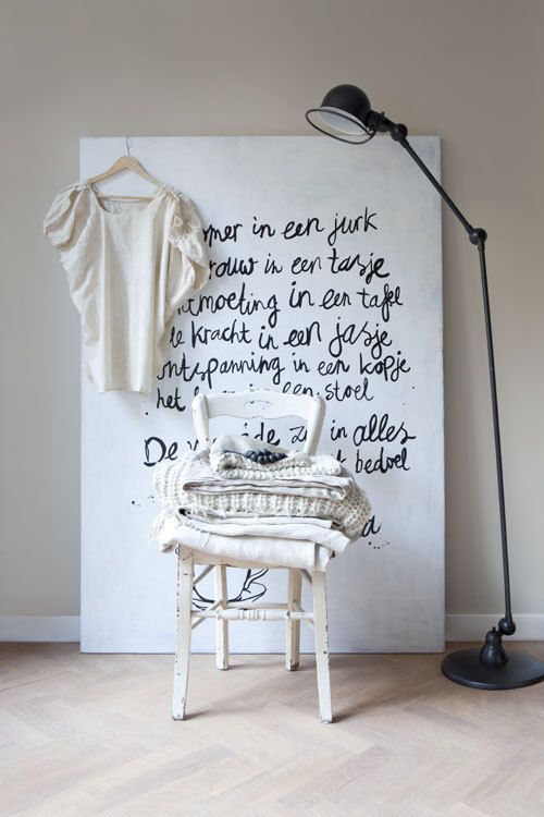 Large white canvas with handwritten black typography in Dutch, styled with a white vintage chair stacked with cozy neutral knitwear, and a floor lamp for a soft minimalist look.