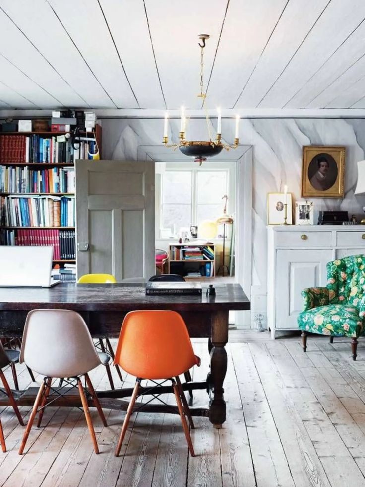 Dining room with distressed white wooden floors and a large dark wood table, surrounded by iconic Eames-style chairs in white, orange, and yellow, illustrating a blend of traditional and Mid-Century Modern design.