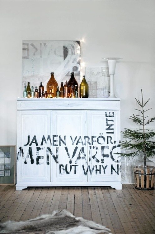 White painted cabinet with large black stenciled typography in Swedish and English, decorated with amber glass bottles on top and a minimal Scandinavian holiday aesthetic.