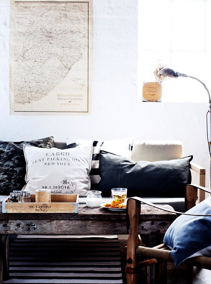 Rustic and cozy living room featuring a rough-hewn wooden coffee table with a wooden tray, a light sofa adorned with mixed-texture pillows (including a large white one with 'CARGO' print), and an antique map above.