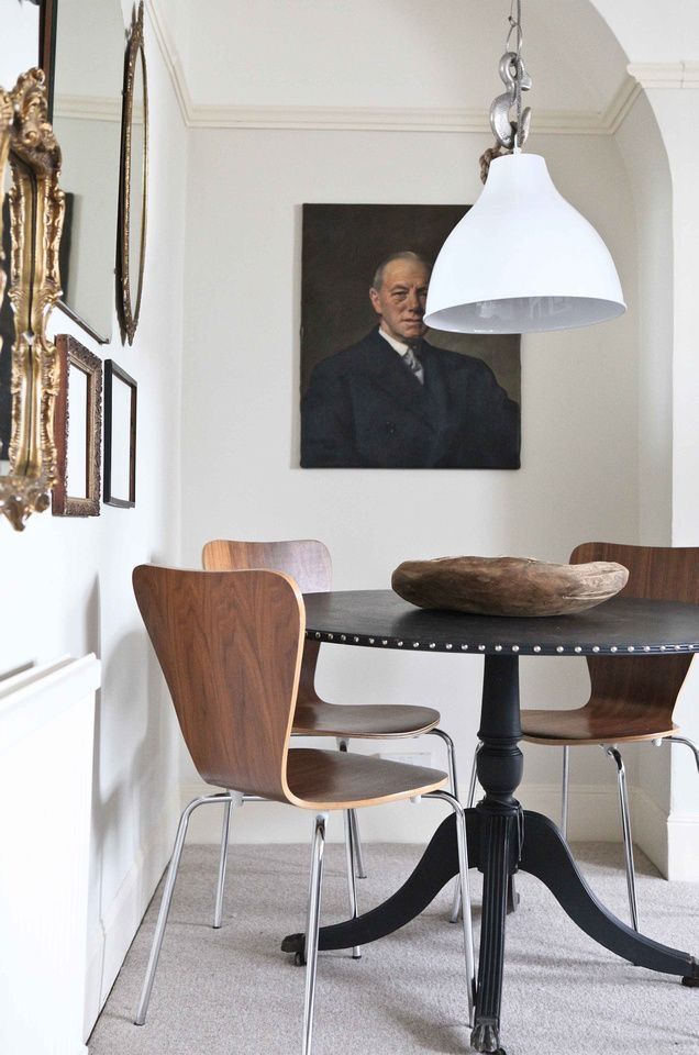An elegant dining area showcasing a blend of traditional and modern elements, with a dark round table and sleek bentwood chairs. A classic portrait hangs on the wall, contrasted by a contemporary white pendant lamp. Decorative gold mirrors add a touch of vintage charm.