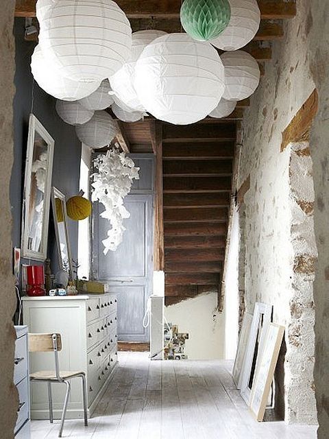 An eclectic hallway featuring a cluster of white and one light green paper lanterns hanging above, with distressed stone walls, exposed wooden ceiling beams, light wood floors, and a white chest of drawers.
