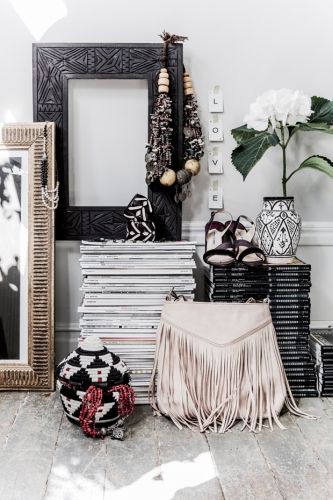 A bohemian-style vignette on a rustic wood floor. A large stack of magazines is topped with a fringed beige handbag, a decorative woven basket, and a carved black picture frame with layered beaded necklaces.