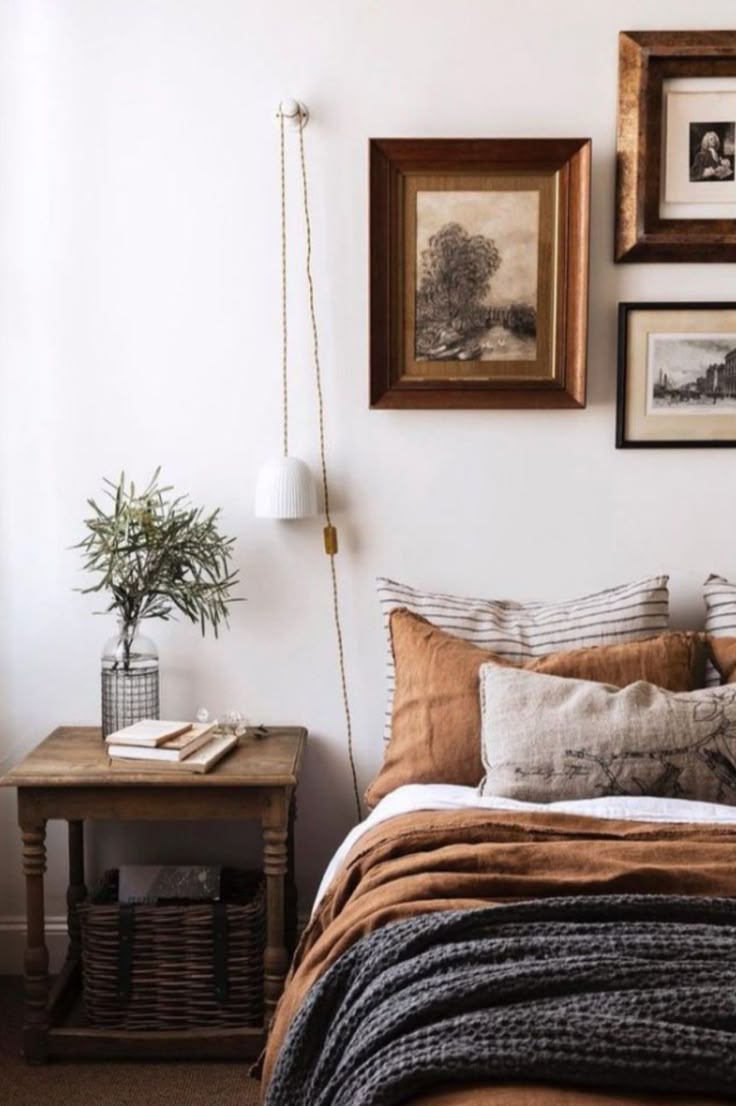 A cozy bedroom with a bed featuring a terracotta-colored linen duvet, a chunky gray throw, and striped pillows. A rustic wood side table holds a glass vase with greenery and is next to a small gallery wall of framed vintage art.