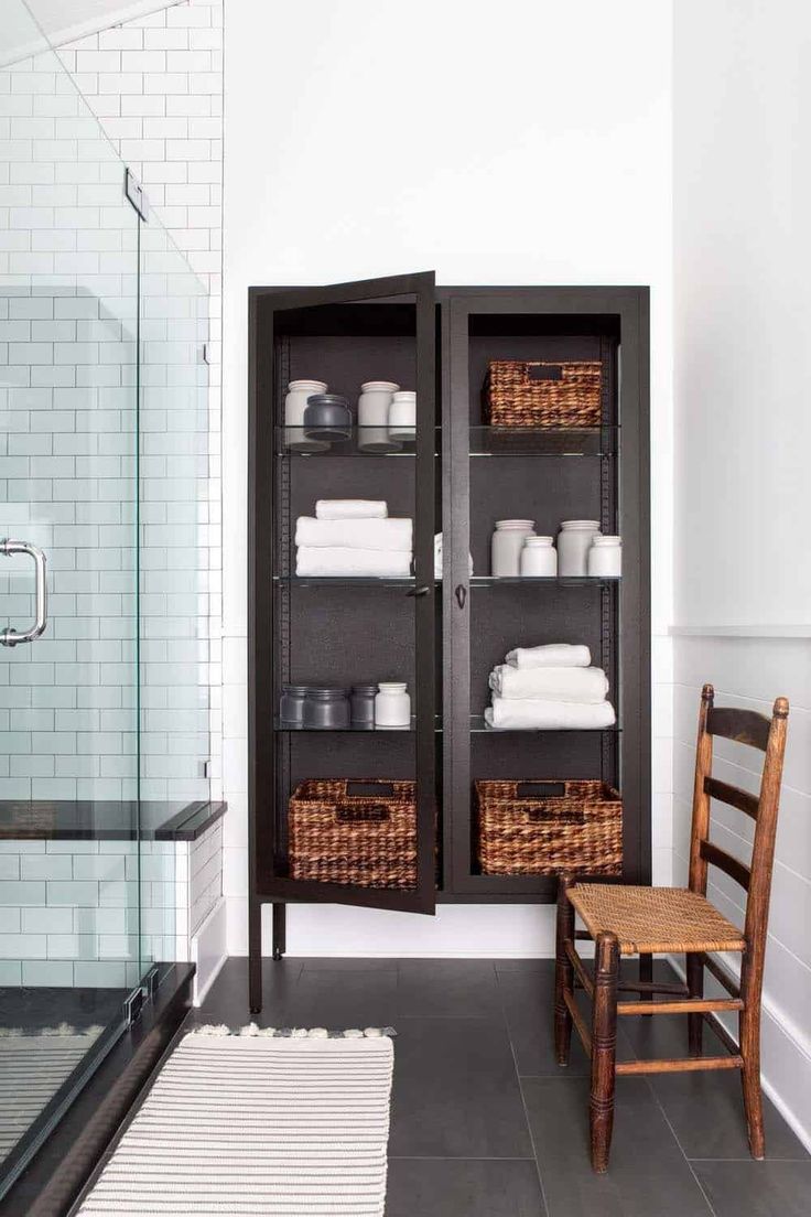 An example of decorating with a bold black cabinet in a bathroom. The black metal storage cabinet is styled with organized towels, lidded jars, and woven baskets, providing both storage and a high-contrast accent in the mostly white space.