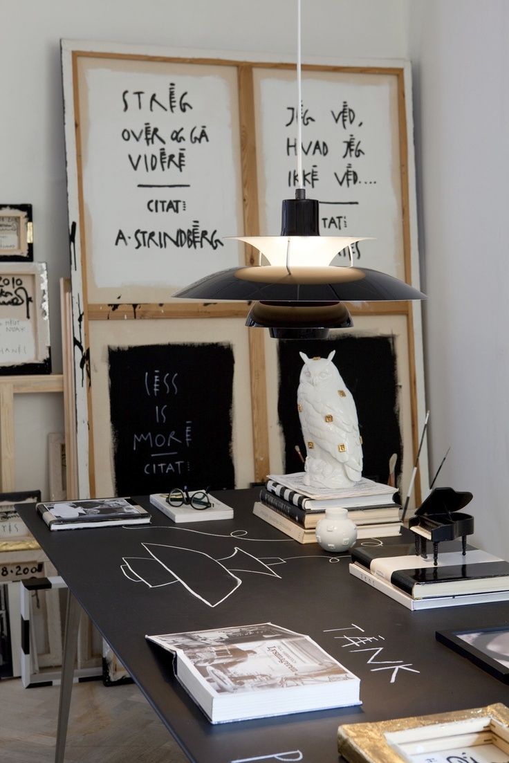 An artistic space with a black table that has chalk drawings on it, holding a stack of books and a white owl figurine. Large canvases with black-and-white writing are in the background.