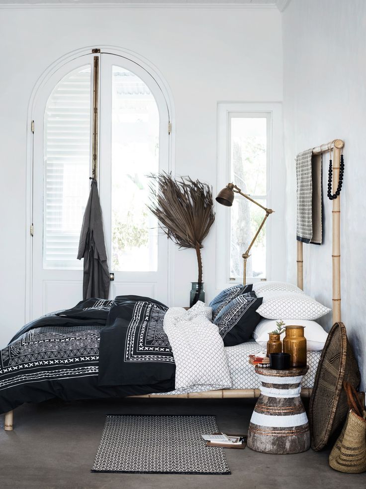 A bohemian-style bedroom featuring a bamboo bed frame with a black and white patterned duvet cover. The space is decorated with a dried palm leaf, a brass lamp, and a side table made from textured, light-colored wood.