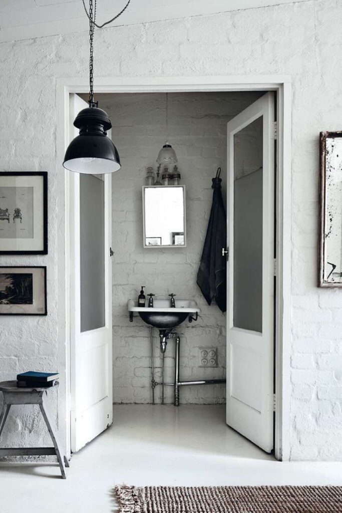 A vintage-style bathroom shown through an open doorway, featuring white exposed brick walls, a small chrome pedestal sink, and a black industrial pendant light hanging in the foreground.