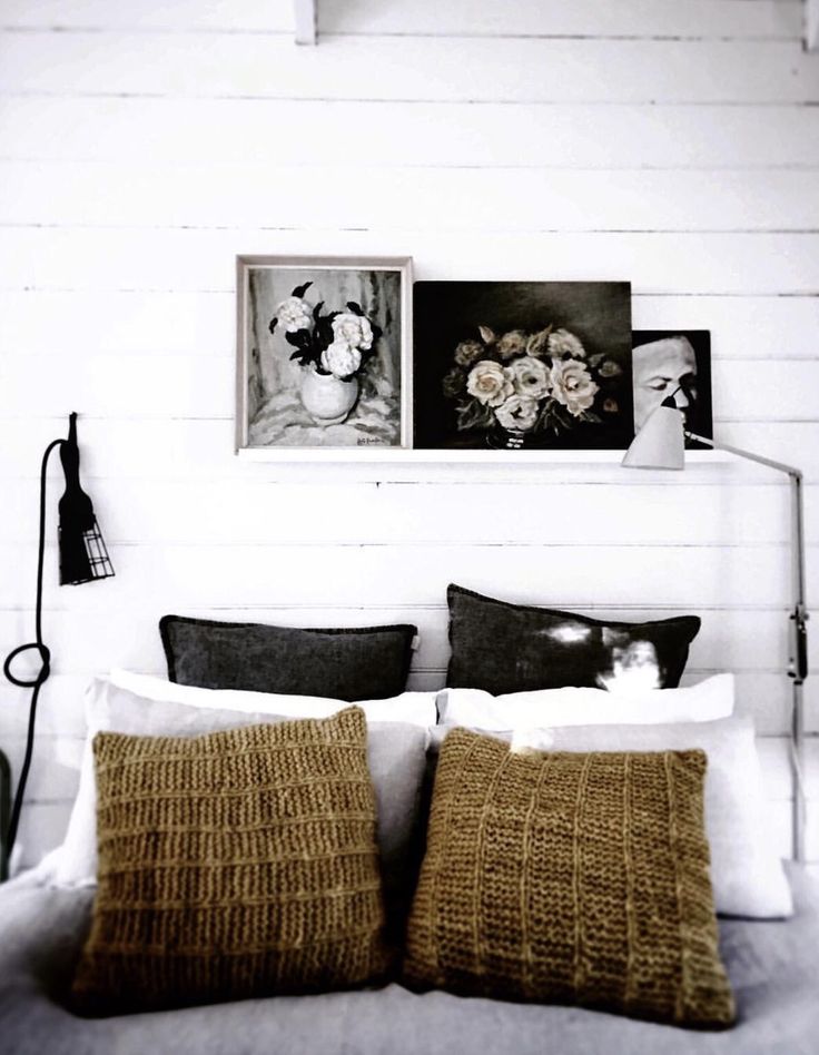 A cozy bedroom with a white paneled wall behind a bed. The bed is styled with a mix of dark gray and chunky knit tan pillows, and a small gallery of floral artwork is propped on a ledge above the headboard.