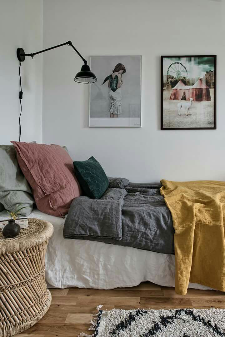 A cozy bedroom featuring a bed with a color-blocked linen duvet and pillows in shades of gray, dusty pink, and mustard yellow. A round rattan side table and an articulating black wall lamp are also visible.