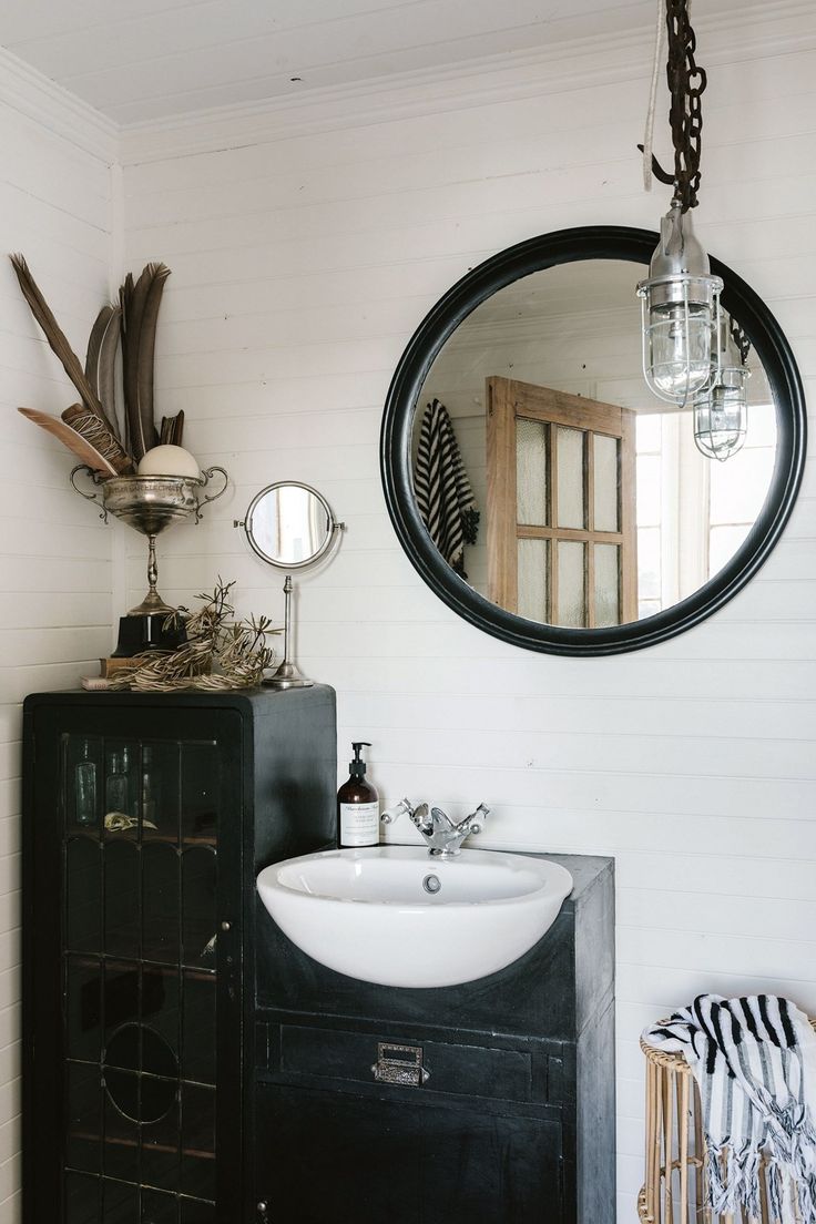 A bathroom with a vessel sink on a rustic black metal cabinet. A large round black-framed mirror hangs above it, and two vintage industrial lights hang from the ceiling.