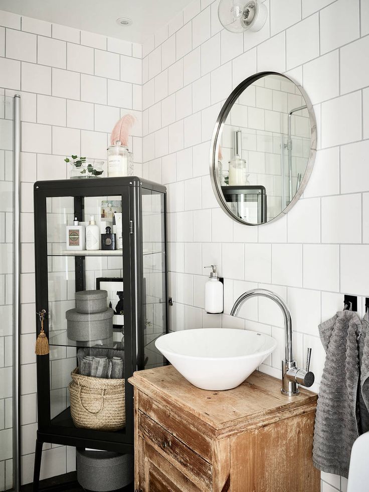 A bathroom featuring white tiled walls, a rustic wood vanity with a white vessel sink, and a black metal and glass storage cabinet next to it. A round, silver-framed mirror hangs above the sink.