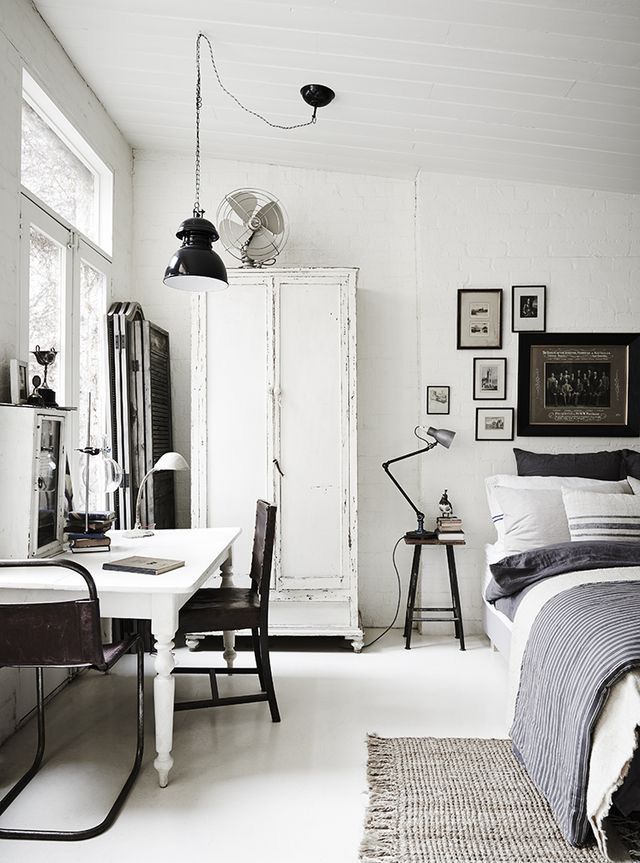 A bright, clean white bedroom and workspace combo. The room features a white desk and a distressed white armoire, with a bed in the background and a gallery wall of framed black-and-white photos.