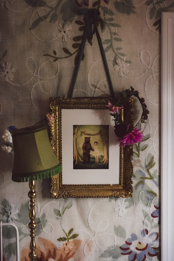 A decorative gold-framed picture of a bear and a person hangs on a wall with vintage floral wallpaper. A green lampshade and a floral wreath are also visible.