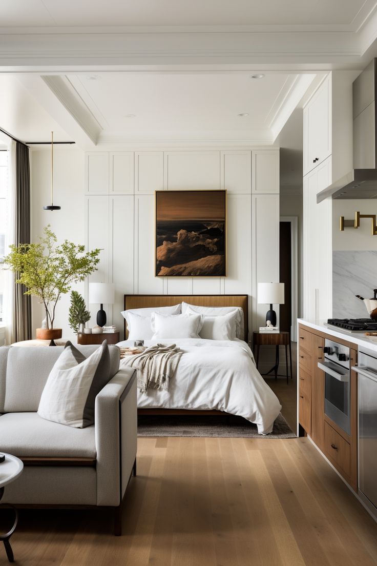 A bright, modern studio apartment with a bed, a sofa, and a kitchen area, all in a light color palette with wood floors.