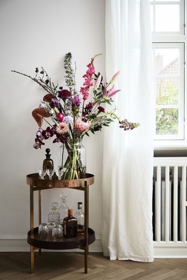 A beautiful, colorful bouquet in a glass vase on a two-tiered bar cart next to a window with sheer curtains.