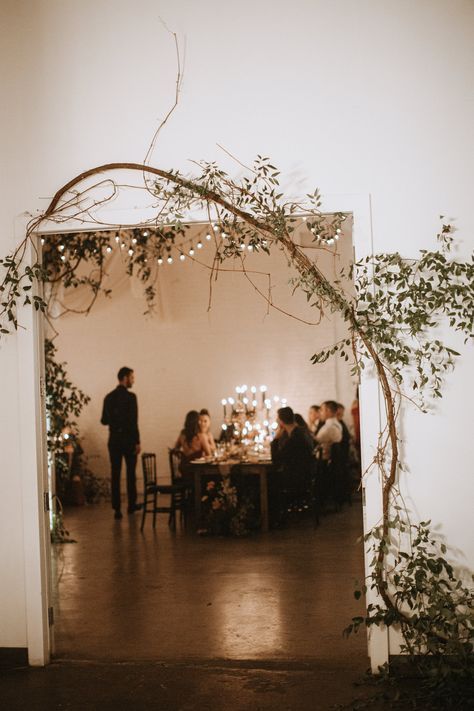 A dining table set with a flowing olive garland used as a centerpiece runner, accompanied by thin white taper candles in brass holders.