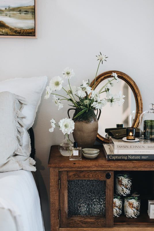 A wooden bedside table featuring a tall ceramic lamp, two hardback books, a small green succulent, and a scented candle, demonstrating the "rule of three" in styling.