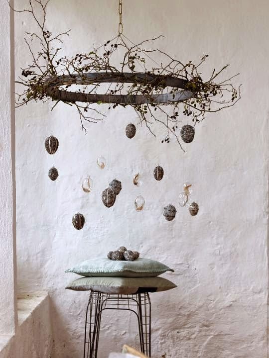 A tall, minimalist Easter egg tree featuring a single architectural bare branch in a matte black ceramic vase. Delicate glass eggs in a uniform champagne hue hang from thin silk threads, reflecting the soft spring light in a modern living room.